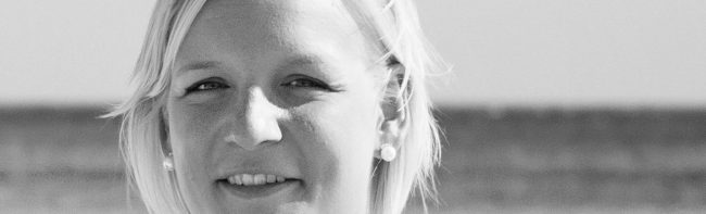 Frau mit blonden Haaren und Ohrringen lächelt in die Kamera, Porträt vor unscharfem Hintergrund.