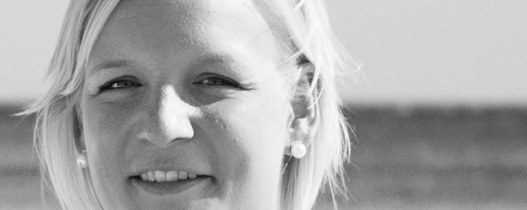 Frau mit blonden Haaren und Ohrringen lächelt in die Kamera, Porträt vor unscharfem Hintergrund.