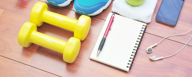 Gelbe Kurzhanteln, Trainingsschuhe, Notizbuch, Stift, Kopfhörer, Handy, Apfel auf Handtuch.