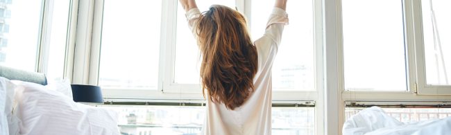 Frau streckt sich im Bett vor einem hellen Fenster, bereit für einen energiegeladenen Tag – perfekt für Fitness- und Ernährungsprogramme.