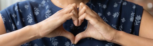 Hände formen Herz vor floralem Oberteil, Symbol für Herzgesundheit und Fitness.