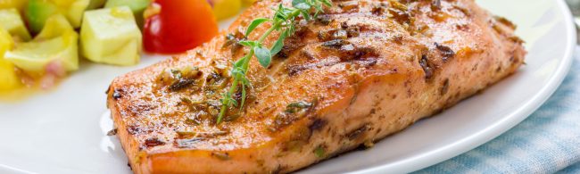 Grilled salmon fillets topped with fresh mango–avocado salsa on a plate, with lime wedges and herbs.