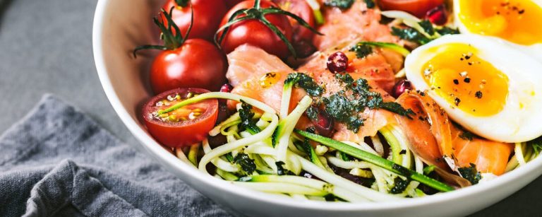 Zucchini noodle salad with smoked salmon, cherry tomatoes and arugula, drizzled with lemon-olive oil dressing on a light plate.