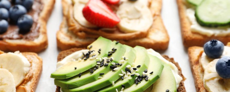 A tray with three healthy sandwich options: a banana‑topped nut butter slice, avocado power toast with seeds, and a Greek‑style sandwich with tzatziki, feta, cucumber, tomato and herbs.