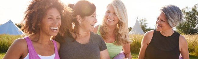 Two friends exercising together outdoors, smiling and doing bodyweight squats side by side in a sunny park.