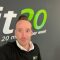 Studiomanager Kees Moët foto fit20 Coach lächelt im Studio, vor dem fit20 Logo. Er bewirbt 20 Minuten effektives Fitness-Training pro Woche.