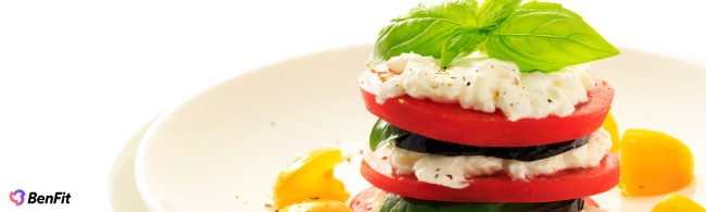 Ein appetitliches, mehrschichtiges Gericht mit frischen Tomaten, cremigem Mozzarella, grünem Basilikum, dunklen Auberginenscheiben und gelber Paprika auf einem weißen Teller. Ideal als gesunde, vegetarische Mahlzeit für eine ausgewogene Fitness-Ernährung von BenFit.
