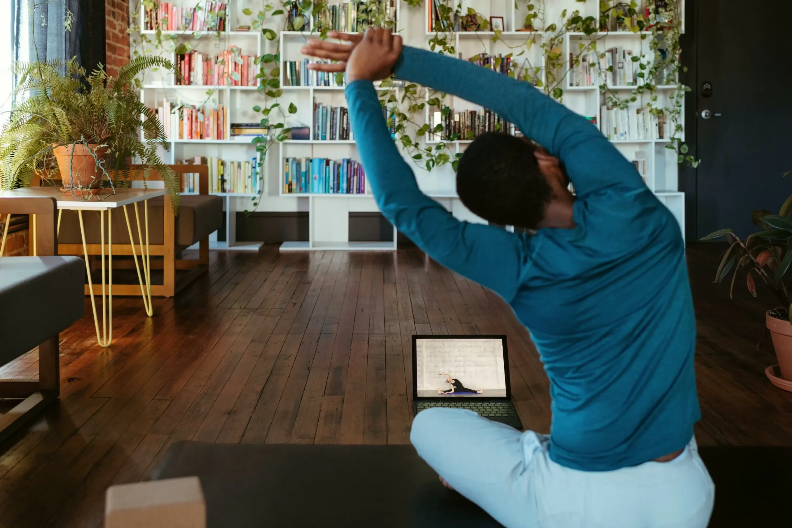 Eine Person macht eine Yoga-Dehnung zu Hause auf einer Matte, folgt einem Online-Fitnesskurs auf einem Laptop. Heimtraining für Flexibilität, Gesundheit und Wohlbefinden in einem inspirierenden Umfeld mit Pflanzen und Büchern.