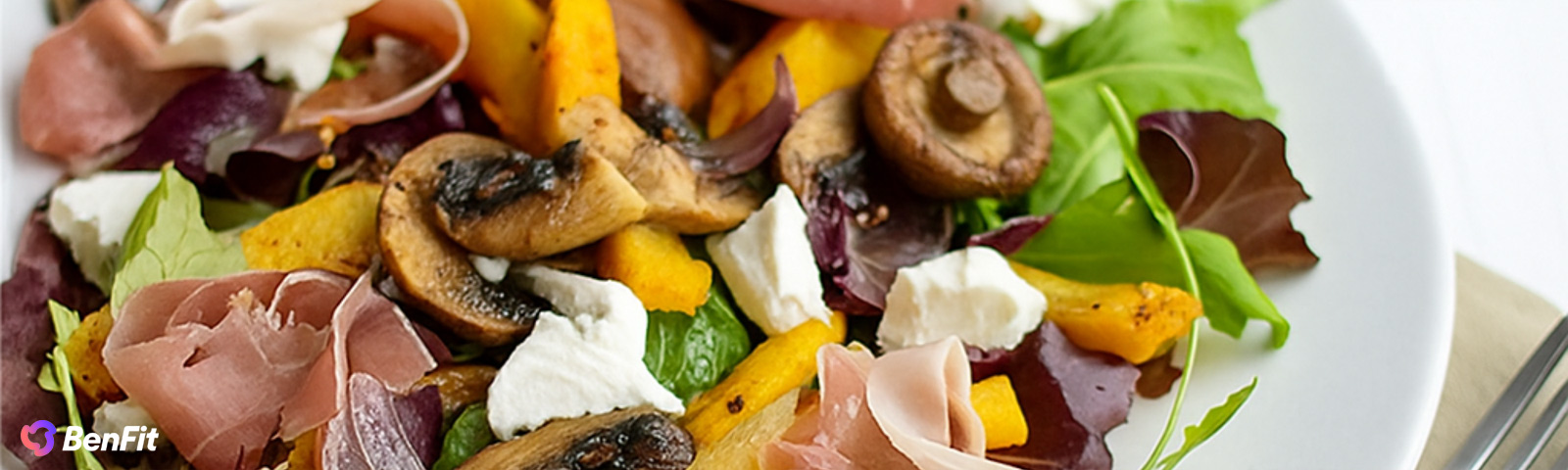 bf_hartverwarmende_herfstalade Nahaufnahme einer gesunden und sausgewogenen Mahlzeit, die zarte sScheiben Schinken, cremigen Ziegenkäse, geröstete Pilze und leuchtend orangefarbenen Kürbis über einem Bett aus frischen grünen und roten Blättern präsentiert.