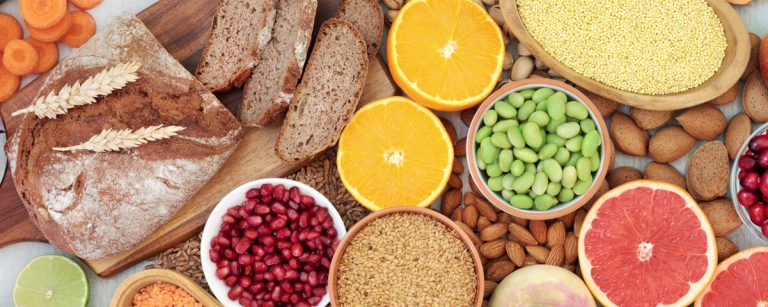 BenFit: Vielfältige Auswahl an gesunden Lebensmitteln wie Vollkornbrot, frischem Obst (Orangen, Grapefruit, Apfel, Cranberries, Granatapfel), knackigem Gemüse (Karotten, Gurken, Zwiebel), Hülsenfrüchten (Linsen, Edamame), Vollkornprodukten (Hirse, Weizen) und Nüssen/Samen (Mandeln, Leinsamen). Ideal für eine ausgewogene, ballaststoffreiche und nährstoffreiche Fitness-Ernährung.